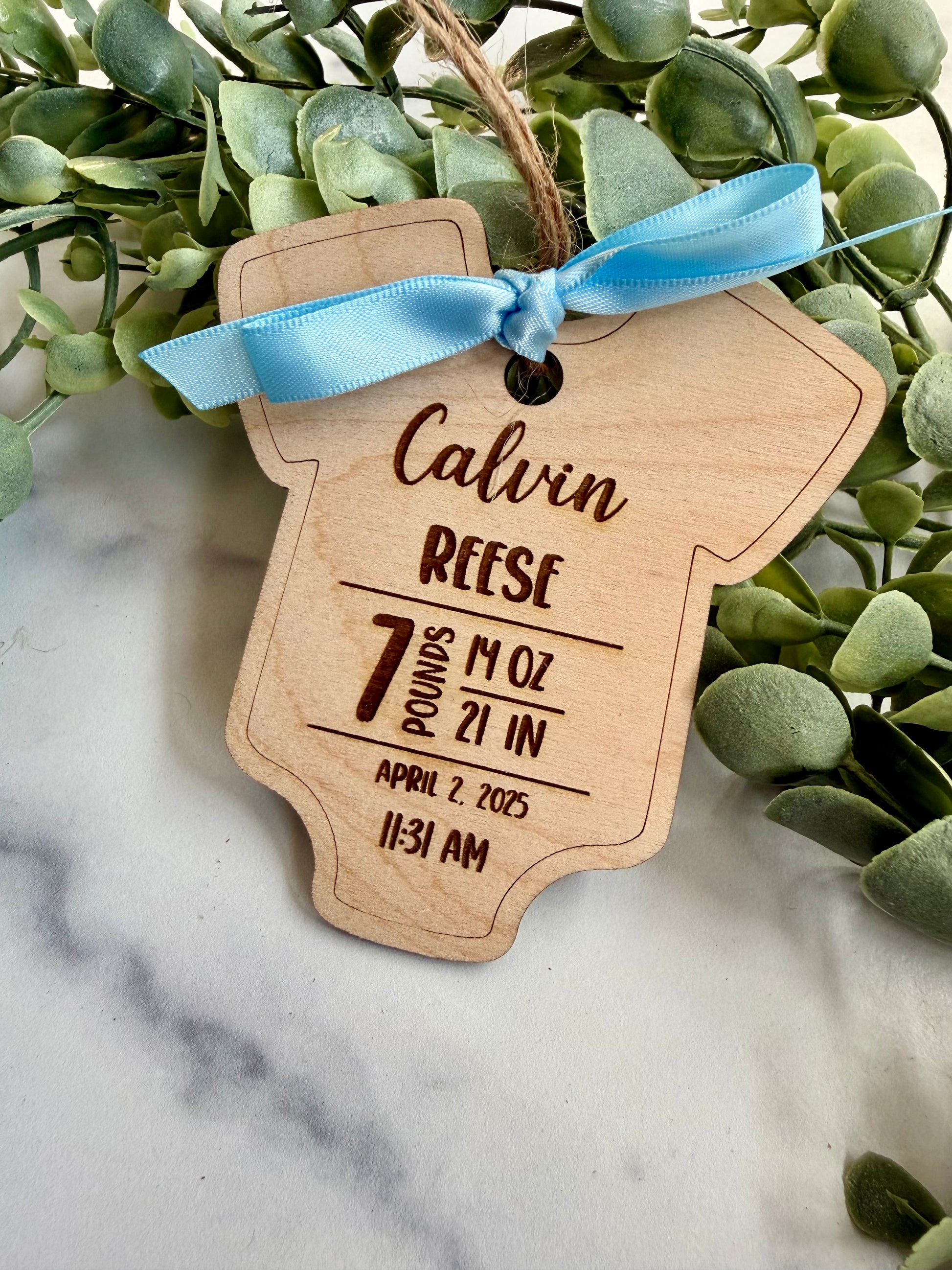 Wooden baby milestone tag with engraved details, tied with a blue ribbon, on a marble surface with green leaves.
