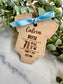 Wooden baby milestone tag with engraved details, tied with a blue ribbon, on a marble surface with green leaves.