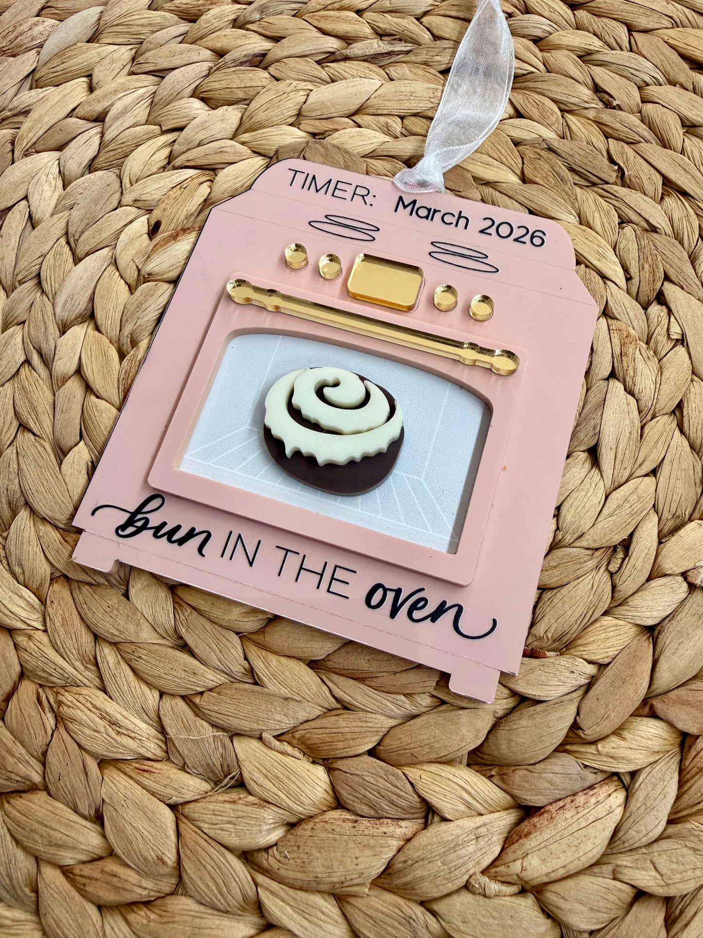 Pink oven-shaped ornament with a bun  and 'bun in the oven' text on a woven surface.
