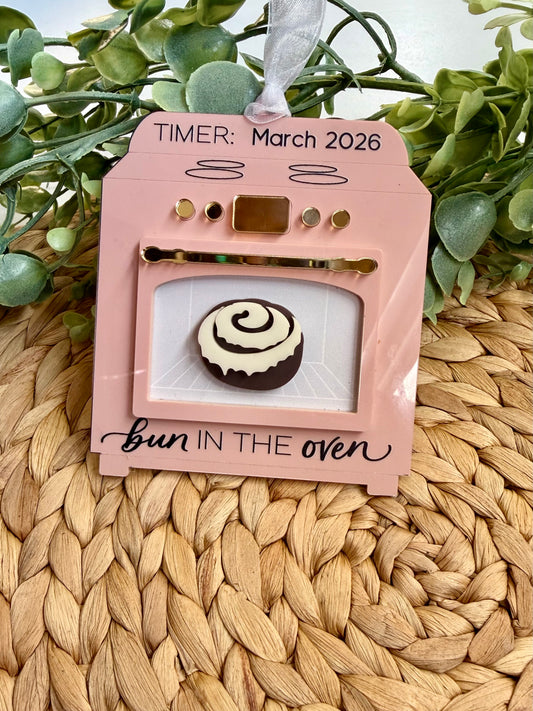 Decorative ornament with one bun in an oven and 'bun in the oven' text on a pink card, set against a woven mat and green leaves background. 