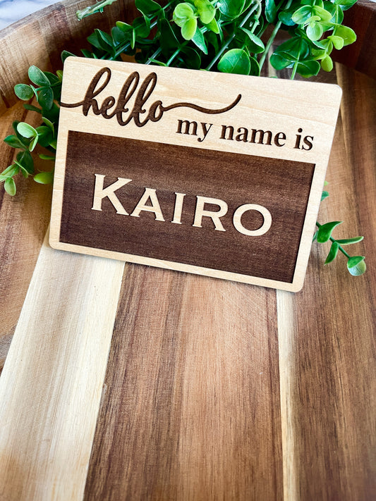 Wooden name tag with 'hello my name is KAIRO' text on a wooden surface with greenery.
