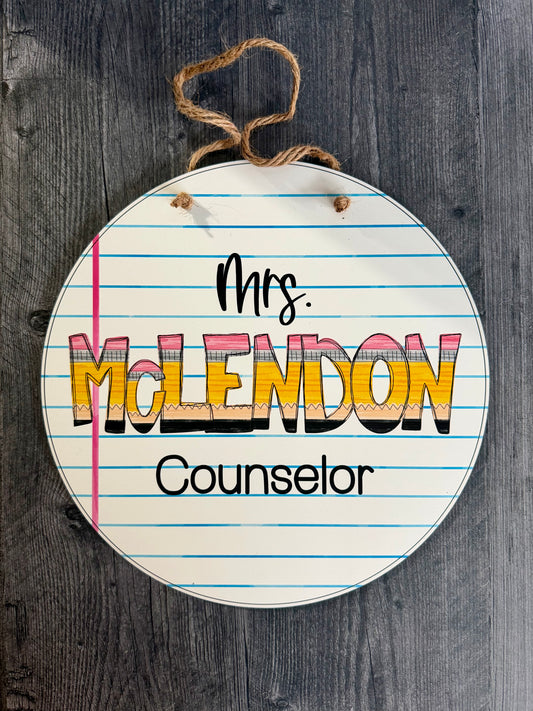 Round sign with lined paper design and 'Mrs. McLendon Counselor' text on a wooden background