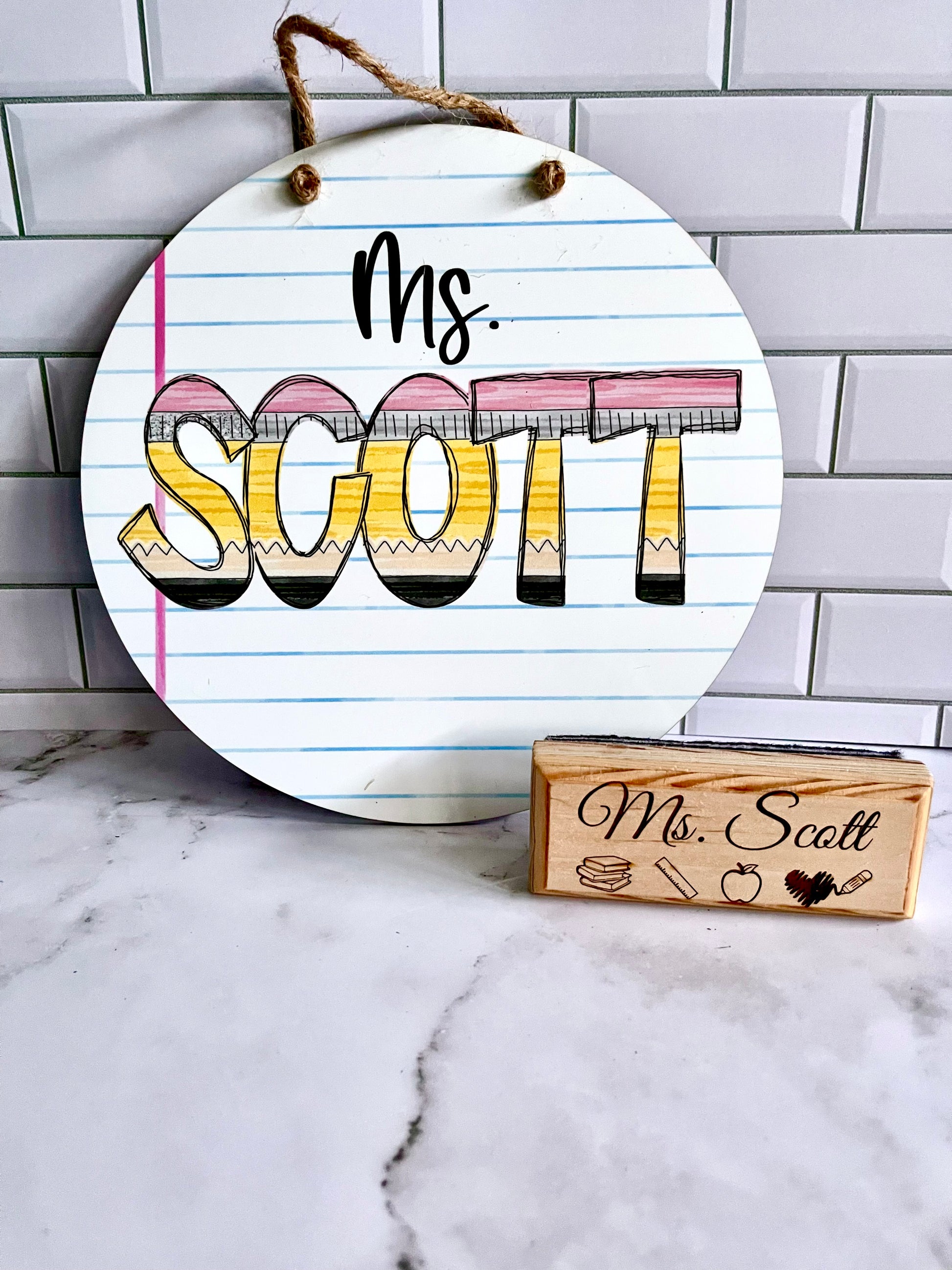 Round wooden sign with 'Ms. Scott' and an eraser on a marble surface and tiled wall background