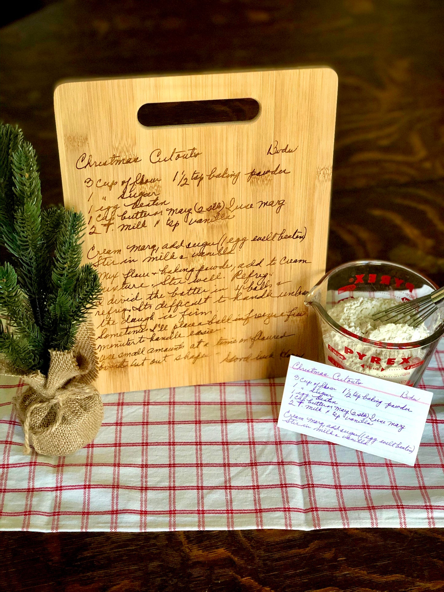 A custom engraved recipe cutting board with handwritten text, displayed next to a small decorative tree and a recipe card, all placed on a checkered tablecloth.