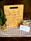 A custom engraved recipe cutting board with handwritten text, displayed next to a small decorative tree and a recipe card, all placed on a checkered tablecloth.