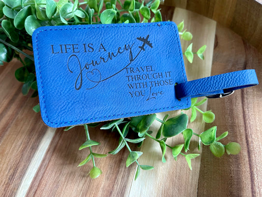 A blue leatherette luggage tag with a silver buckle and black text that reads 'Life is a Journey, Travel with those you love'. It is displayed on a wooden surface with green plant leaves around it.