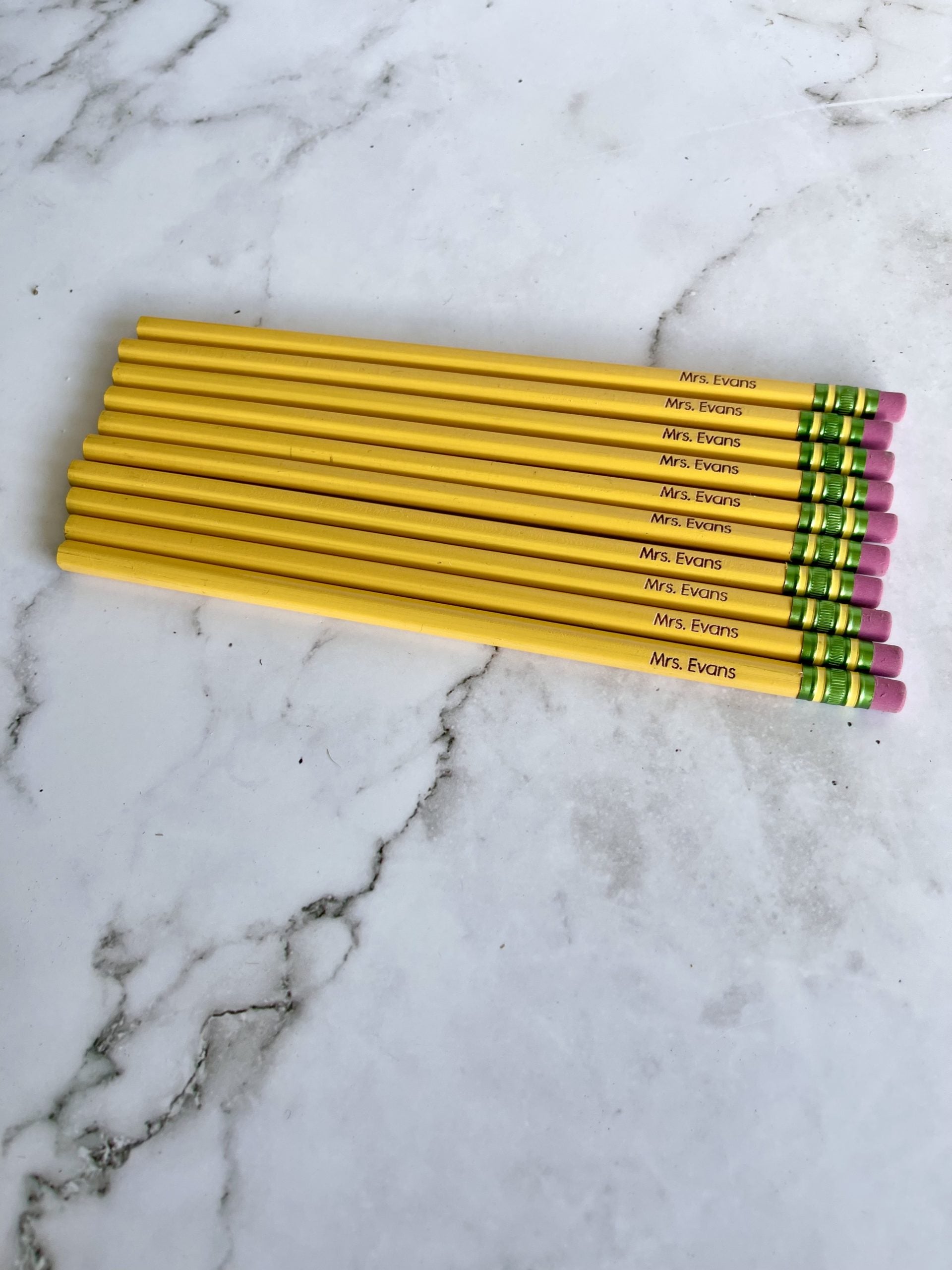A set of yellow Ticonderoga pencils with orange erasers personalized with a name, placed on a marble surface.