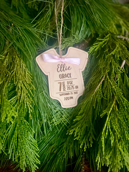 A wooden ornament in the shape of a baby onesie with personalized birth statistics engraved on it, hanging on a Christmas tree.
